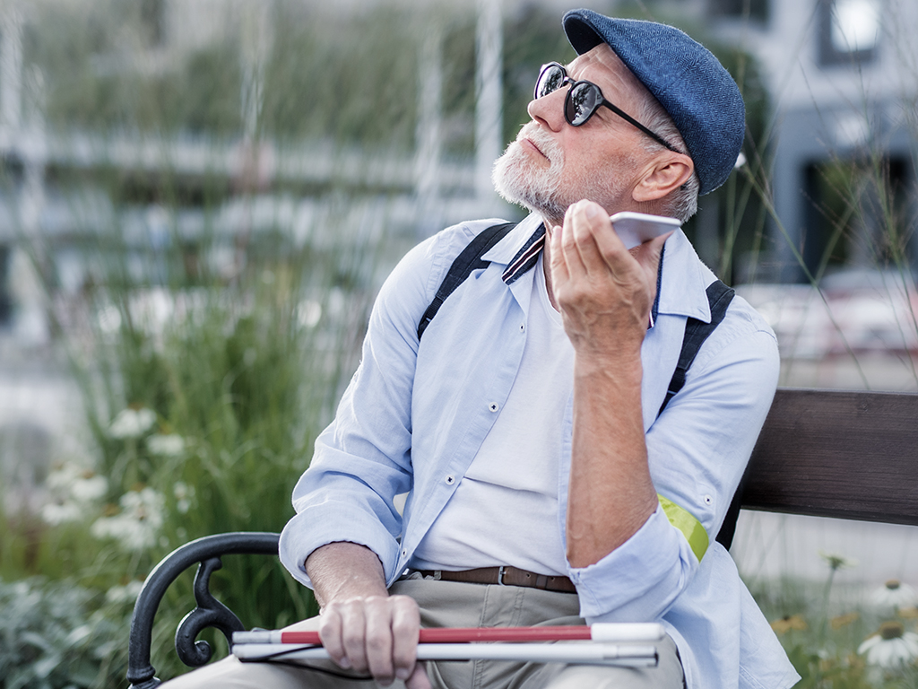 Ein alter, blindet Mann mit Blindenstock sitzt auf einer Bank und nutzt die Smartphone-Sprachausgabe