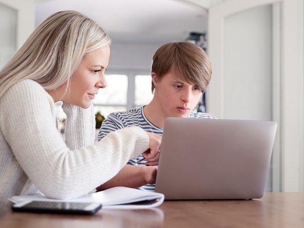 Junge Mutter sitzt zusammen mit einem Teenager mit Down-Syndrom am Computer