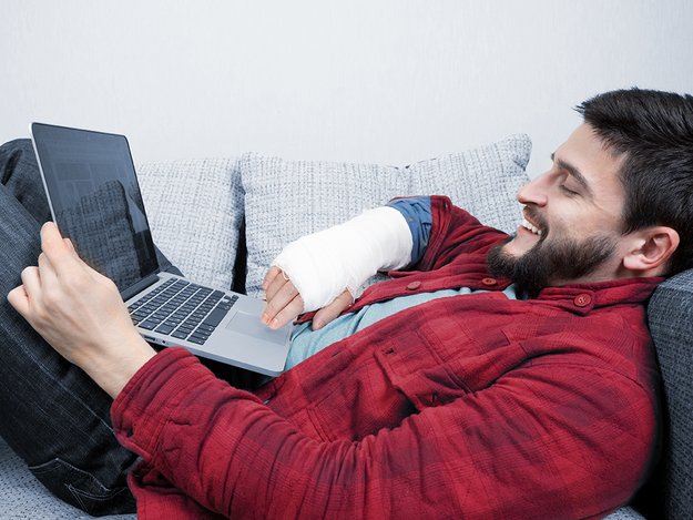 Behinderung kann temporär sein. Ein junger Mann mit Gipsarm liegt auf dem Rücken, ein Laptop auf dem Bauch. Er nutzt die Tastatur zur Bedienung.