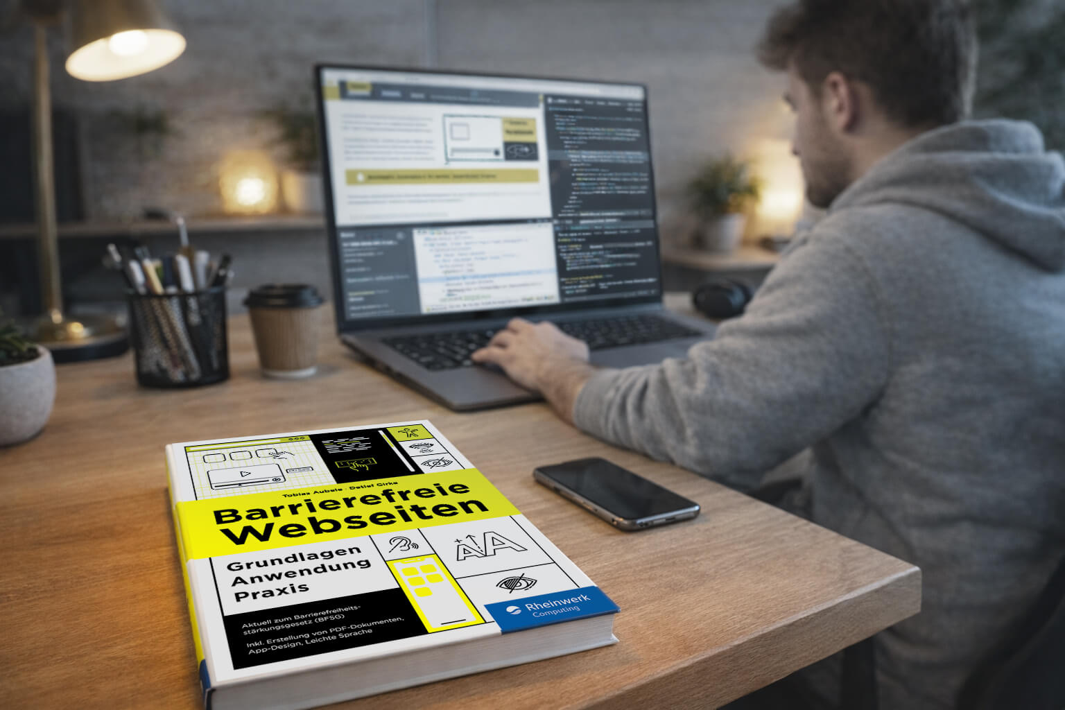 Großansicht Das Buch „Barrierefreie Webseiten – Grundlagen, Anwendung, Praxis“ liegt im Vordergrund auf einem Schreibtisch, im Hintergrund ist ein Mann zu sehen, der an einem Laptop eine Webseite programmiert.
