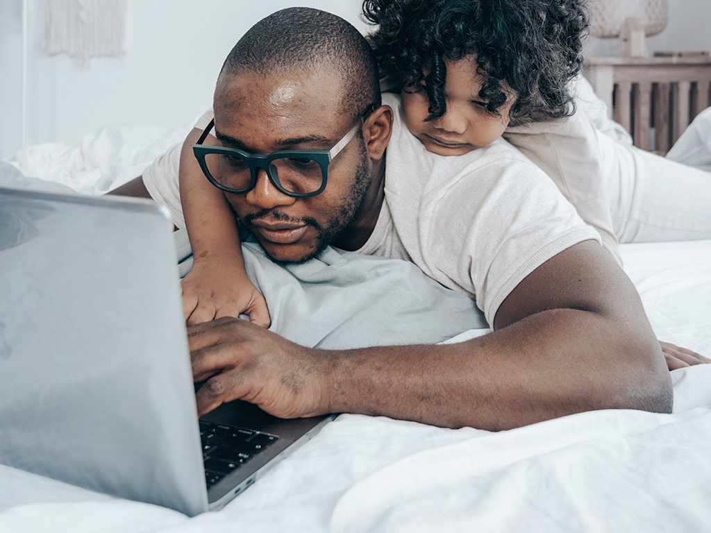 Ein dunkelhäutiger Mann mit Brille liegt im Bett und arbeitet am Laptop. Auf ihm klettert sein Kind herum